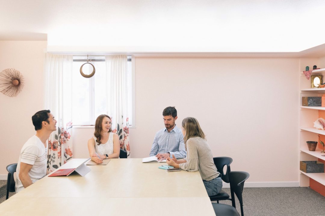 Photo of four people sitting around a conference table inside the Cohere conference room. The walls are light pink and everyone is smiling and laughing.